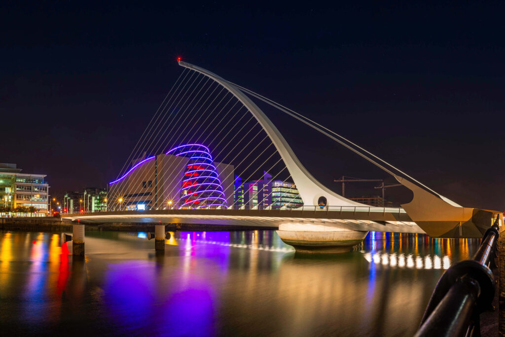 The harp-shaped Samuel Beckett Bridge lit in purple and blue, reflected in the River Liffey at night